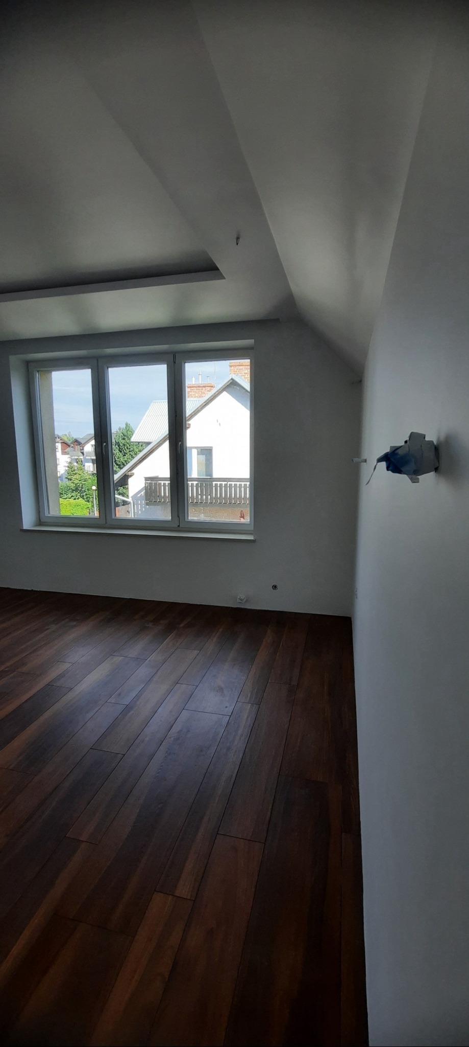 Wnętrze pokoju na poddaszu z drewnianą podłogą, świeżo pomalowanymi na biało ścianami i oknem z widokiem na domy.