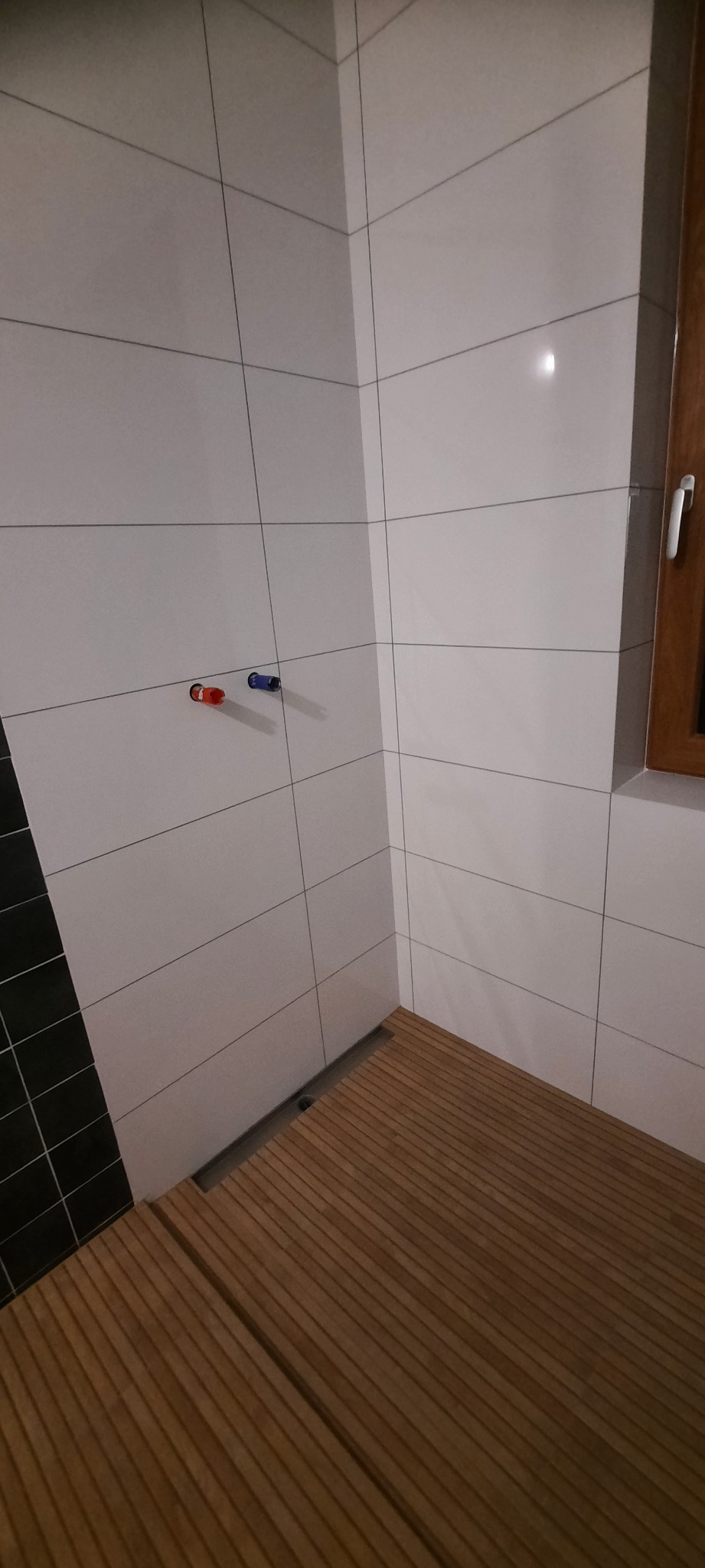 Nowoczesna łazienka po remoncie: białe, prostokątne płytki na ścianach, drewniana podłoga z odpływem liniowym, widoczne przyłącza wody (czerwone i niebieskie) oraz fragment czarnej ściany z płytek.
