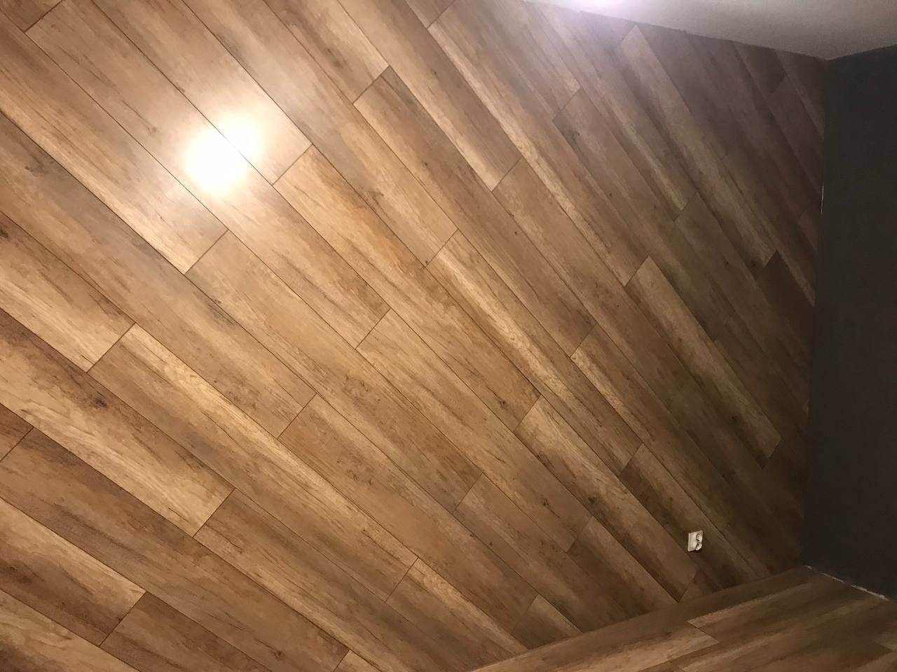 Ukośnie ułożone panele podłogowe imitujące drewno na ścianie w rogu pomieszczenia, widoczny fragment czarnej ściany i gniazdko elektryczne.