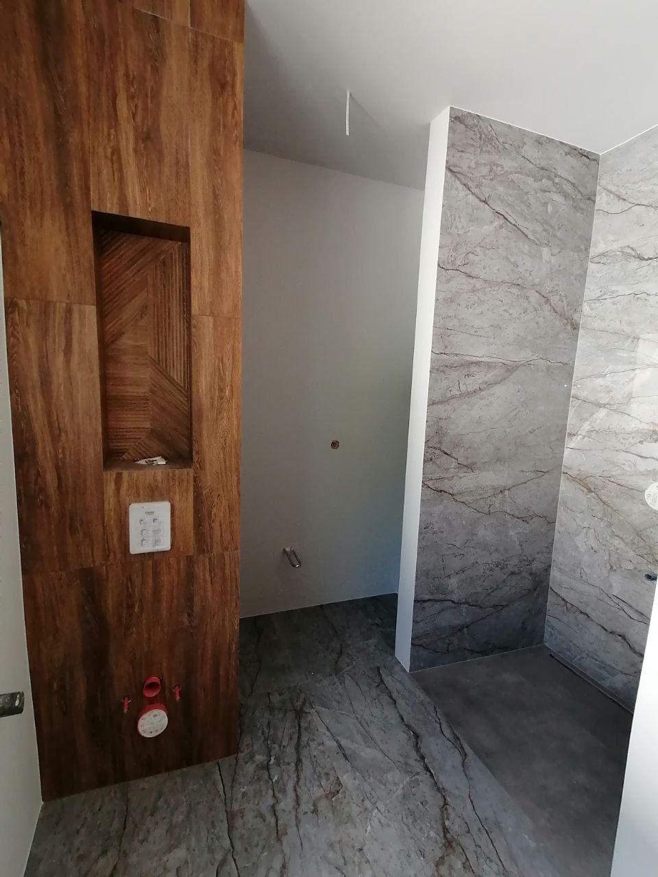 Wykończona łazienka z płytkami imitującymi drewno i marmur; ściana z wnęką i ukrytym systemem spłukiwania, szare płytki podłogowe z wzorem marmuru.