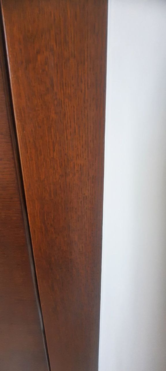 Fragment drewnianej ramy okiennej lub drzwiowej w kolorze ciemnego orzecha, przylegającej do białej ściany. Widoczna struktura drewna.