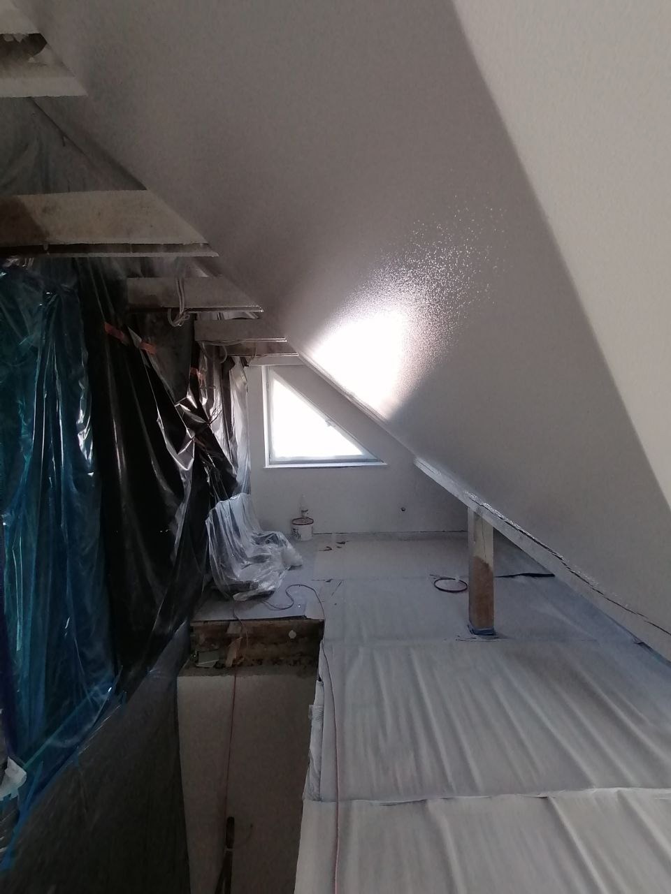 Poddasze w trakcie prac wykończeniowych: pomalowany sufit, zabezpieczona folią podłoga, widoczne drewniane elementy konstrukcyjne i okno dachowe.