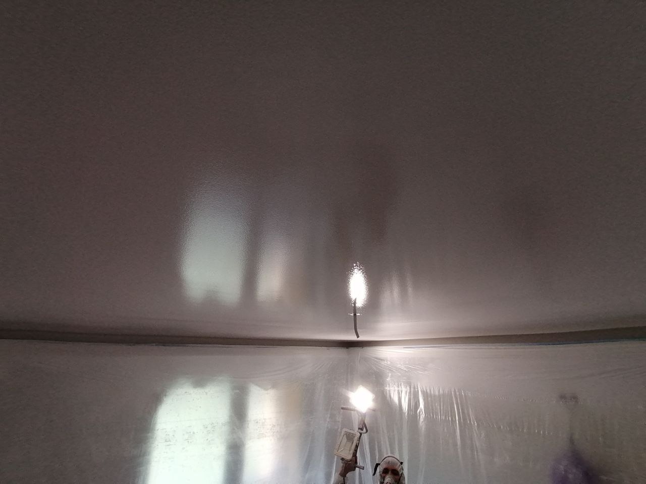 Świeżo pomalowany sufit w pomieszczeniu zabezpieczonym folią malarską, widoczne lampy oświetlające powierzchnię i fragment sylwetki malarza w masce ochronnej.