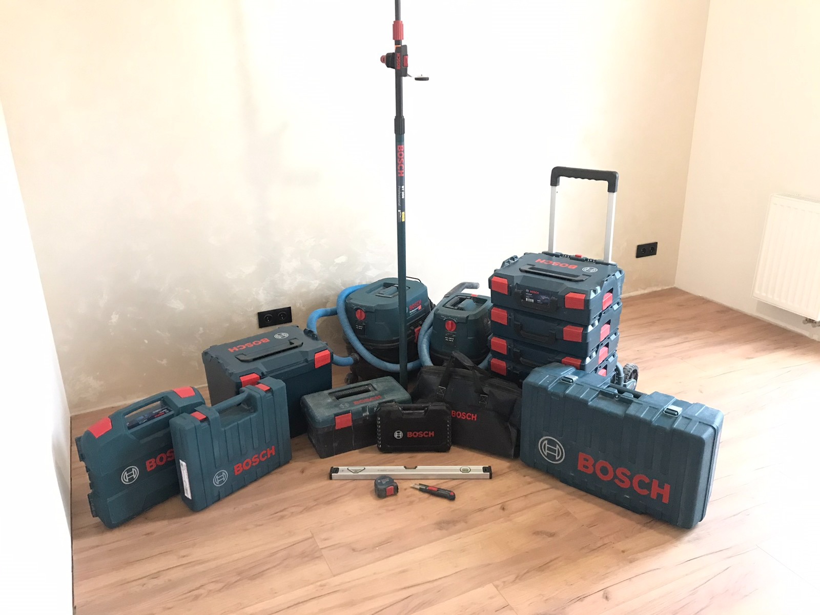 Zestaw narzędzi i akcesoriów marki Bosch, w tym skrzynki narzędziowe, odkurzacze przemysłowe i niwelator laserowy na statywie, rozłożone na podłodze z paneli w pomieszczeniu z surowymi ścianami...