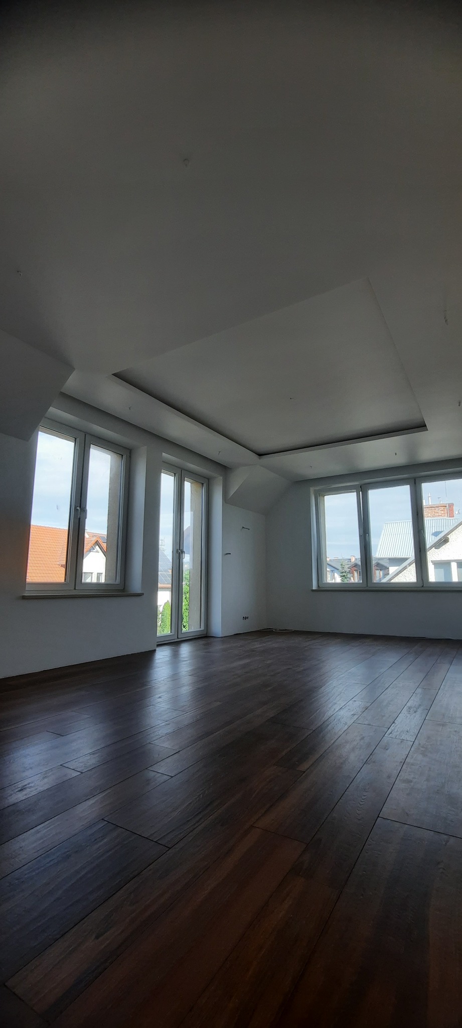 Puste, jasne poddasze z ciemną drewnianą podłogą i białym sufitem z dekoracyjną wnęką, widok na zewnątrz przez okna.