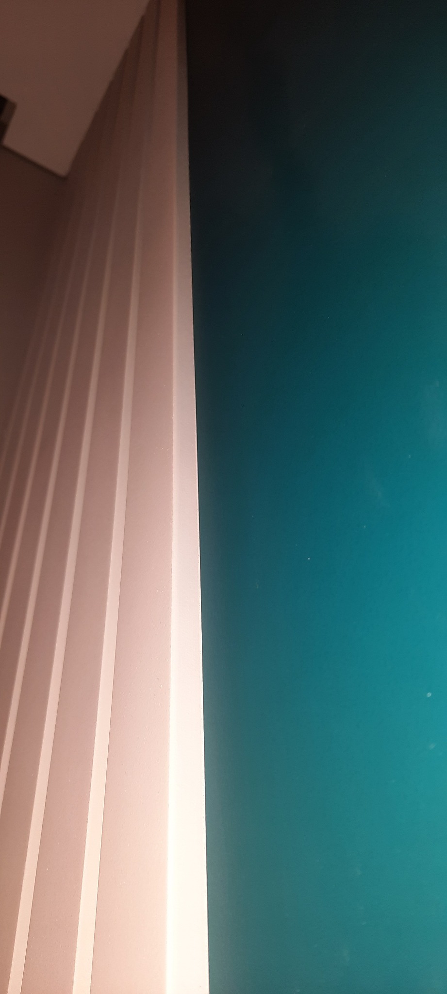 Fragment ściany pomalowanej na intensywny turkusowy kolor, sąsiadującej z dekoracyjną, pionową boazerią w odcieniach bieli i beżu, widzianej pod kątem.