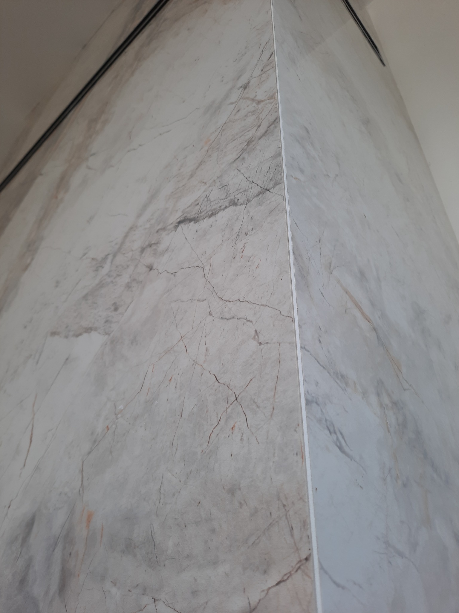 Wewnętrzny narożnik wykończony płytami imitującymi marmur, z widoczną listwą narożną i fragmentem sufitu z czarną linią wentylacyjną.