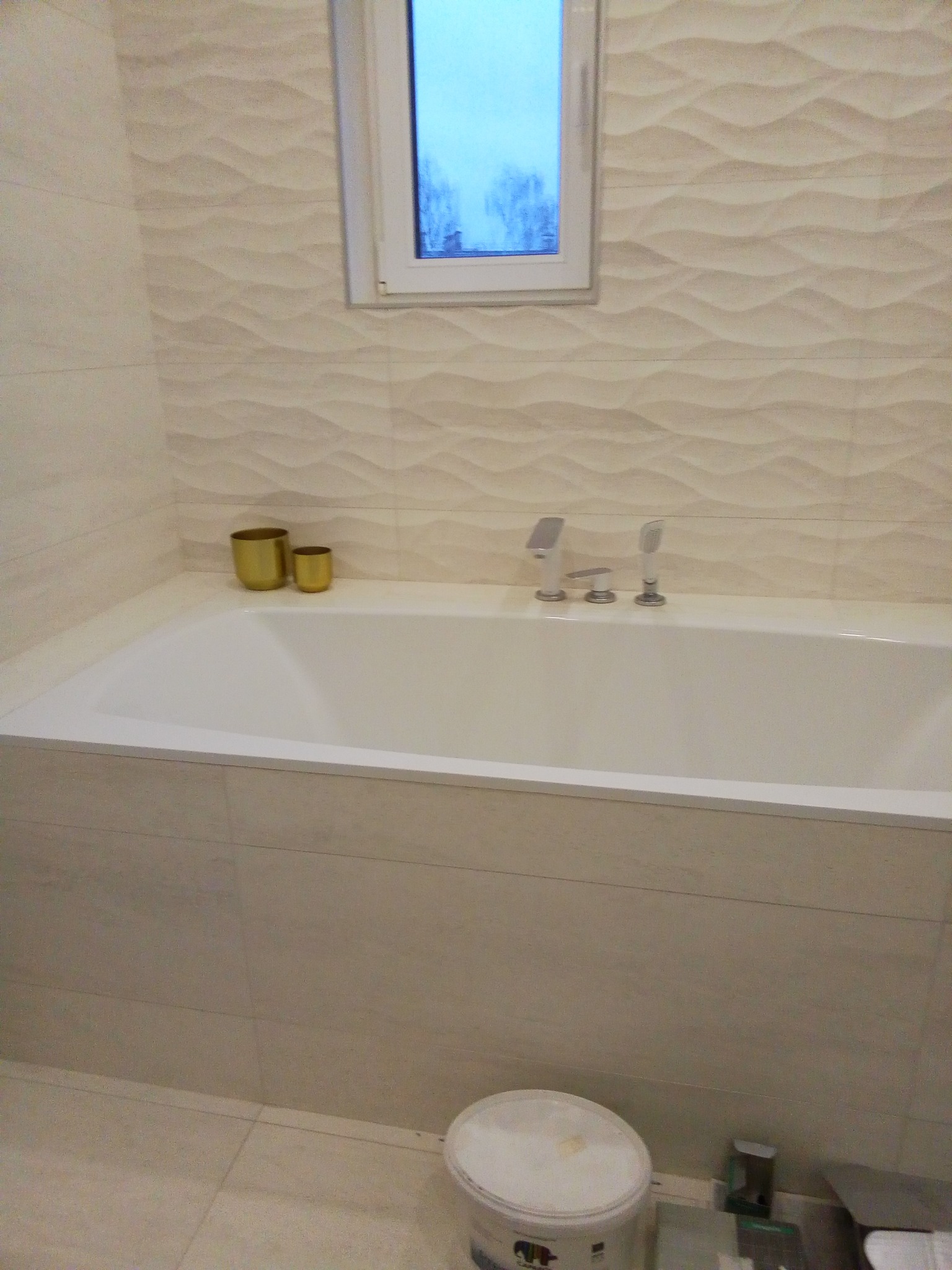 Wykończona łazienka z wanną obudowaną płytkami, ściany wyłożone płytkami z delikatnym wzorem fal, okno z widokiem na drzewa, widoczny kubeł z farbą i narzędzia malarskie przy podłodze.