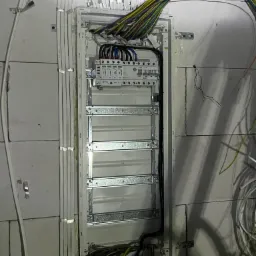 Rozdzielnica elektryczna w trakcie montażu na ścianie z betonu komórkowego; widoczne kable w peszlach i przewody wielokolorowe.