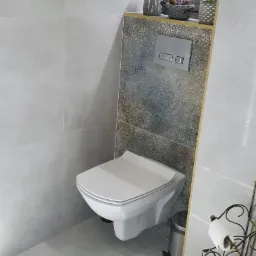 Nowoczesna toaleta podwieszana z szarą płytką imitującą kamień, dekoracyjną półką z podświetleniem LED, na której stoją sukulenty i figurka, oraz metalowym stojakiem na papier toaletowy.