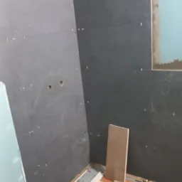 Ściany łazienki w trakcie układania ciemnych, matowych płytek. Widoczne krzyżyki dystansowe, wywiercone otwory na instalacje oraz wiadro z zaprawą i narzędzia na podłodze.