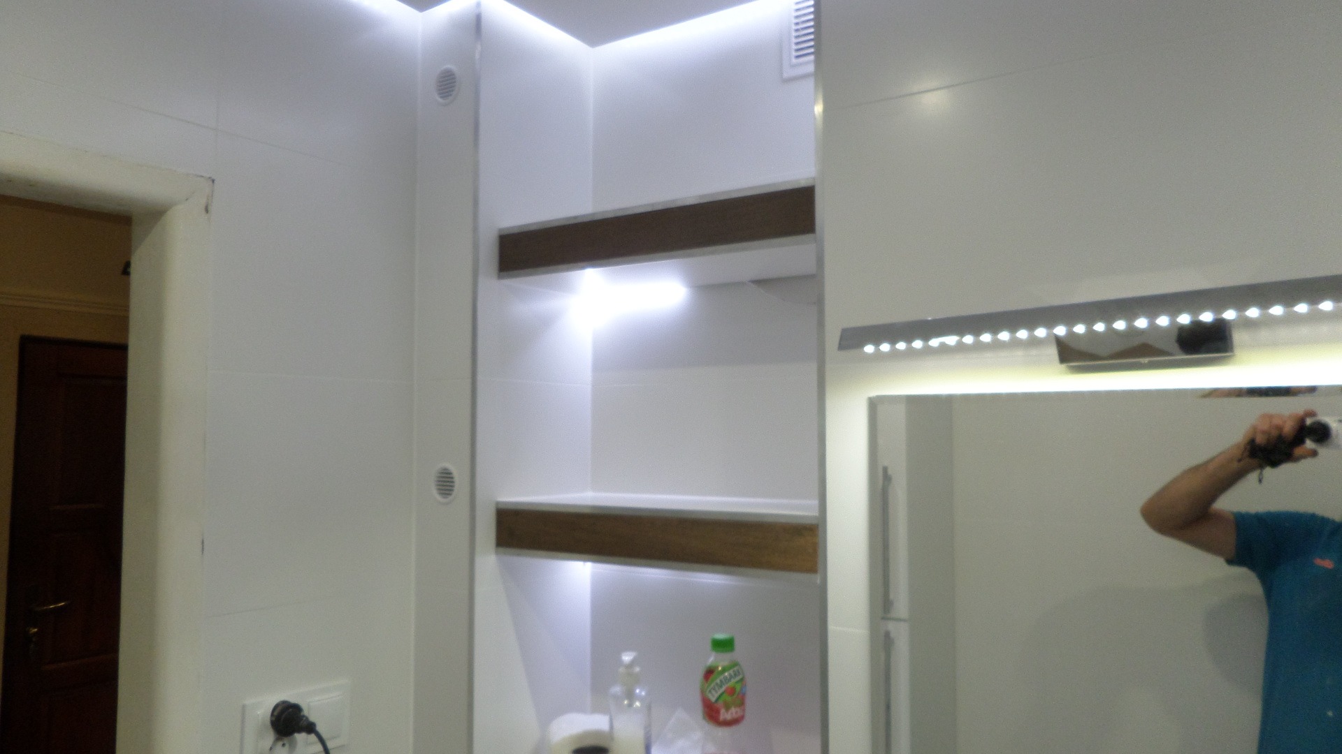 Nowoczesna, jasna łazienka z białymi płytkami, podświetlanymi półkami z drewnianymi elementami i dużym lustrem z oświetleniem LED, z widocznym odbiciem osoby robiącej zdjęcie.