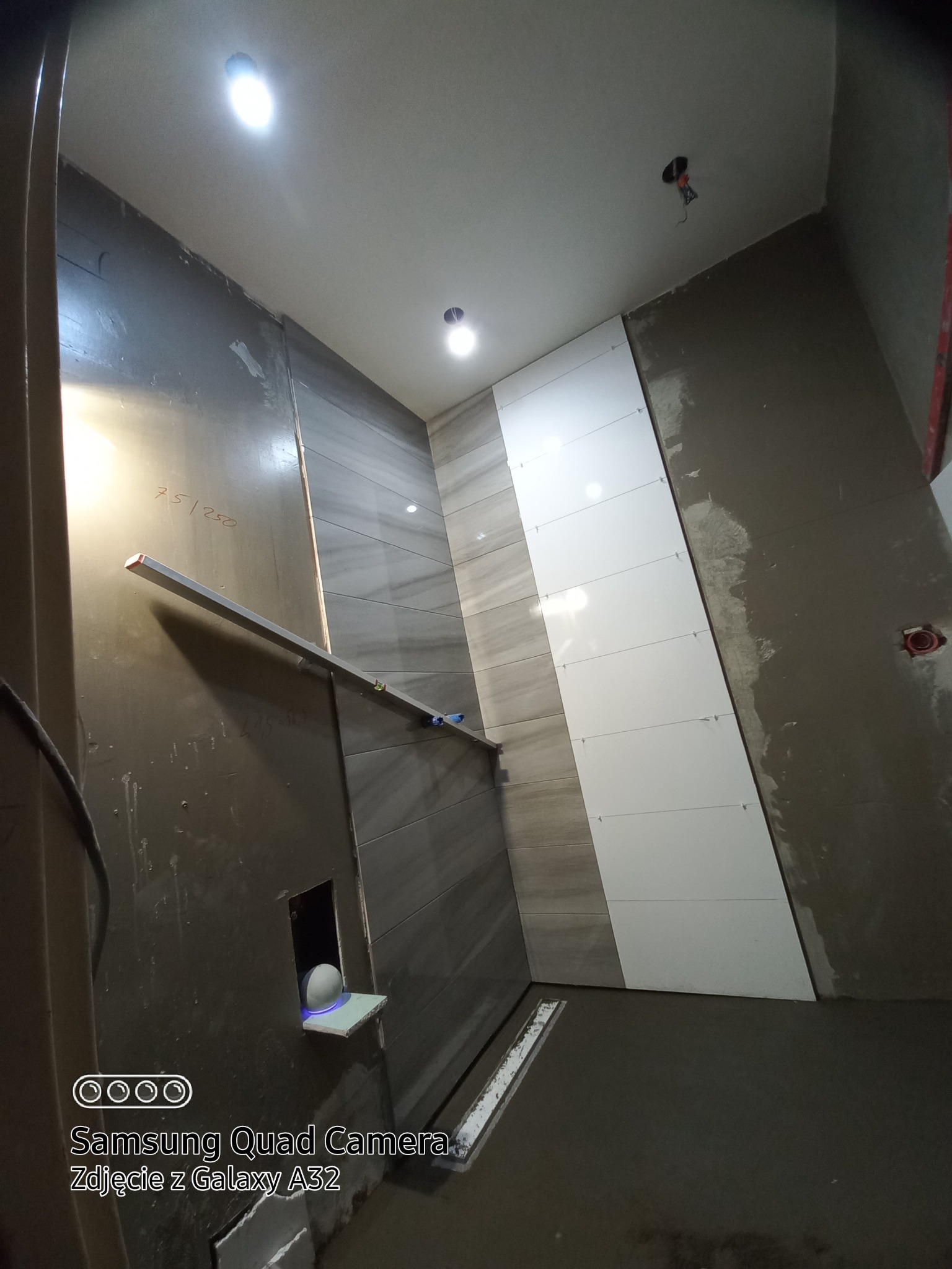 Świeżo ułożone, biało-szare płytki na ścianie łazienki w trakcie remontu, z widocznymi krzyżykami dystansowymi i poziomnicą, surowy stan podłogi i ścian.