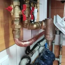 Instalacja hydrauliczna z miedzianymi rurami, zaworami z czerwonymi uchwytami i filtrami wody z ciemnymi obudowami, zamontowana na ścianie z panelami drewnopodobnymi.