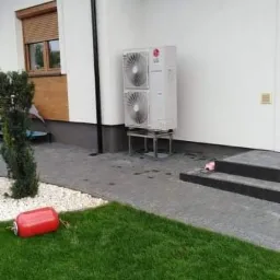 Biała jednostka zewnętrzna pompy ciepła LG, zamontowana na metalowym stelażu przy białej ścianie domu z czarną dachówką, widok na trawnik i czerwony zbiornik.