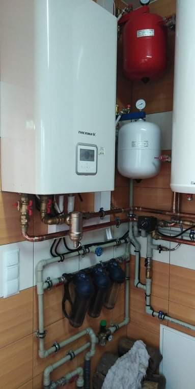 Biały piec grzewczy Thermav z wyświetlaczem, połączony miedzianymi rurami z czerwonym zbiornikiem wyrównawczym i białym zbiornikiem ciśnieniowym, dodatkowo widoczne filtry wody i instalacja wodna...