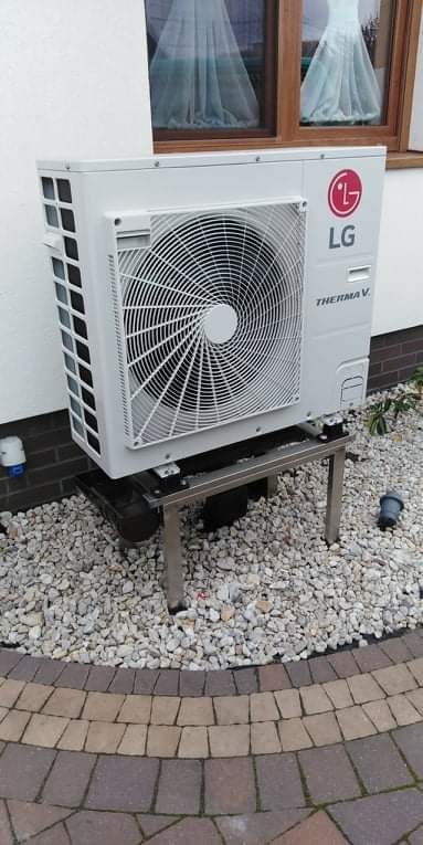 Biała zewnętrzna jednostka pompy ciepła LG Therma V umieszczona na metalowym stelażu, otoczona białymi kamykami i kostką brukową, z oknem w tle.