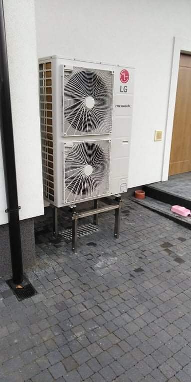Zewnętrzna jednostka pompy ciepła LG Therma V, zamontowana na metalowym stelażu na kostce brukowej, z widoczną rurą spustową przy białej ścianie budynku.