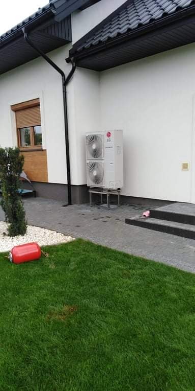 Biała jednostka zewnętrzna pompy ciepła LG, zamontowana na metalowym stelażu przy białej ścianie domu z czarną dachówką, widok na trawnik i czerwony zbiornik.