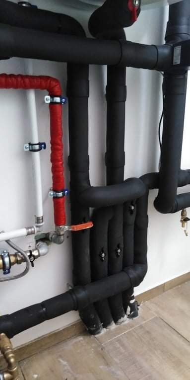 Różnokolorowe rury grzewcze, czarne zaizolowane i czerwone, przymocowane do białej ściany z widocznymi zaworami i przyłączami.