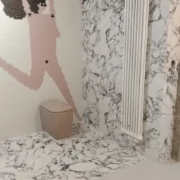 Nowoczesna łazienka z marmurowymi płytkami na ścianach i podłodze, mozaika z motywem postaci na ścianie obok toalety, biały grzejnik.