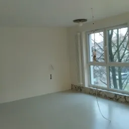 Pusty, świeżo wyremontowany pokój z białymi ścianami, grzejnikami i trzema oknami z widokiem na drzewa bez liści. Na ścianie widoczne ślady po instalacjach elektrycznych.