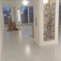 Wnętrze jasnego, przestronnego pokoju z szarą podłogą, widoczne okna, grzejnik, dekoracyjny filar z marmurową wnęką i kartony na podłodze, prawdopodobnie w trakcie remontu lub wykończenia.