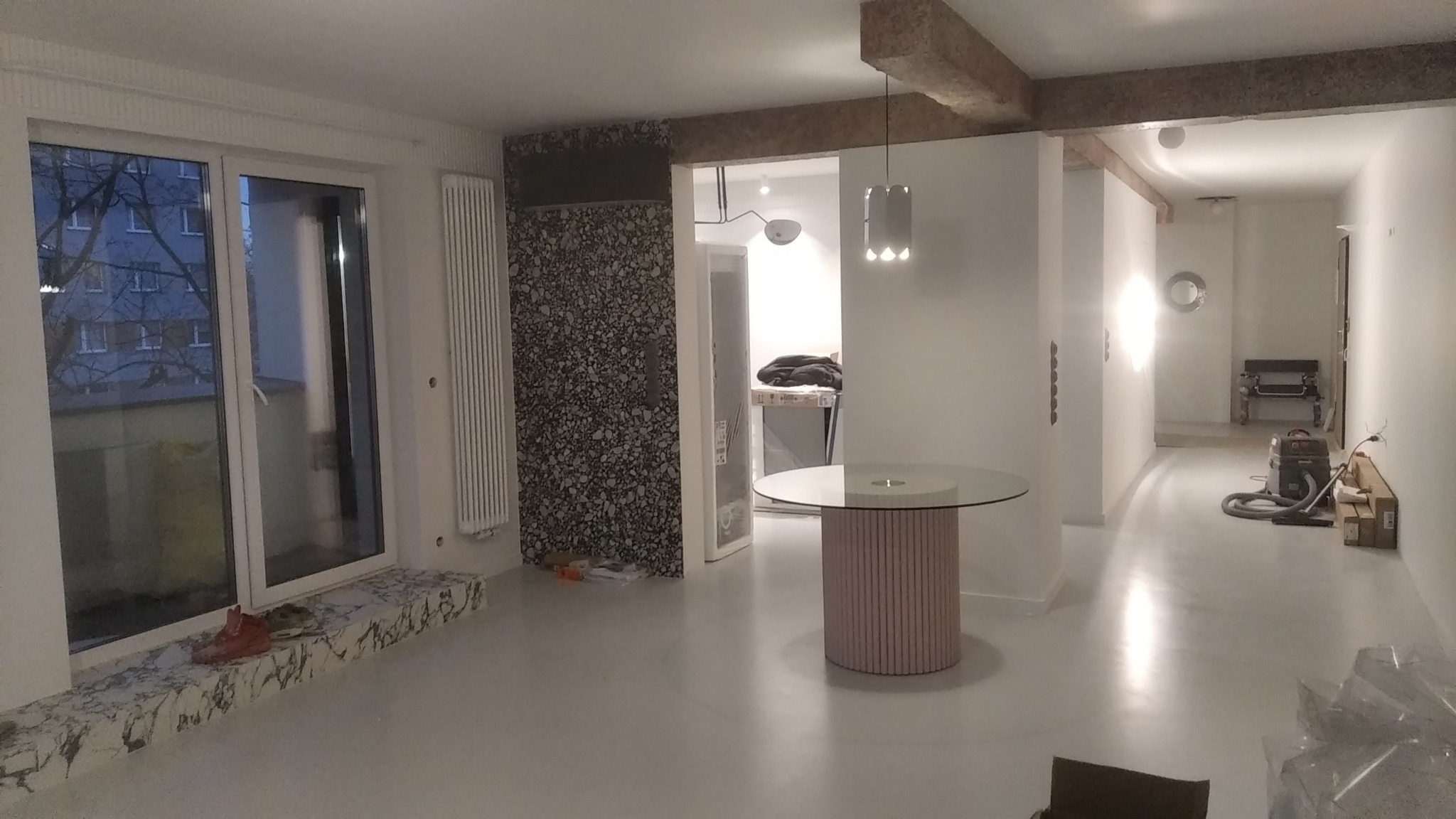 Wnętrze w trakcie wykańczania: białe ściany, szare podłogi, okno z marmurowym parapetem, fragment dekoracyjnej ściany i surowe belki stropowe, widoczny odkurzacz przemysłowy w głębi
