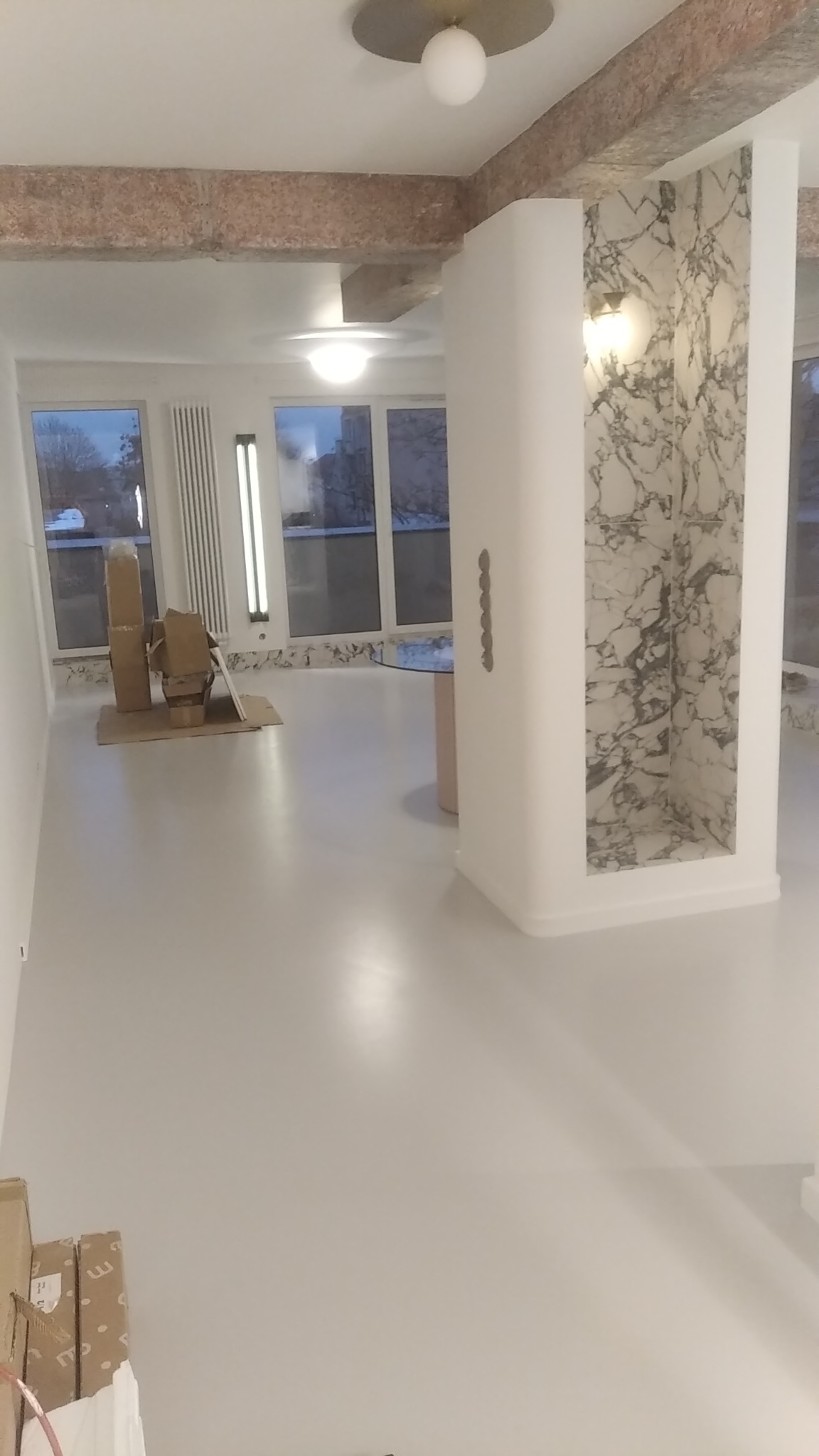 Wnętrze jasnego, przestronnego pokoju z szarą podłogą, widoczne okna, grzejnik, dekoracyjny filar z marmurową wnęką i kartony na podłodze, prawdopodobnie w trakcie remontu lub wykończenia.