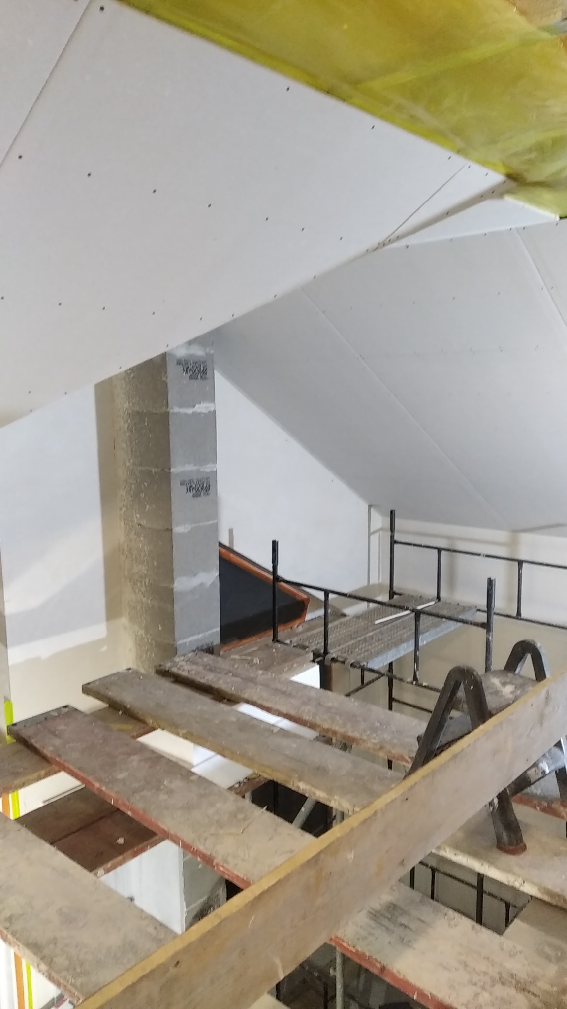 Skośne poddasze z widoczną izolacją i częściowo zamontowanymi płytami gipsowo-kartonowymi, rusztowanie robocze z drewnianymi deskami i fragment komina z betonu komórkowego.
