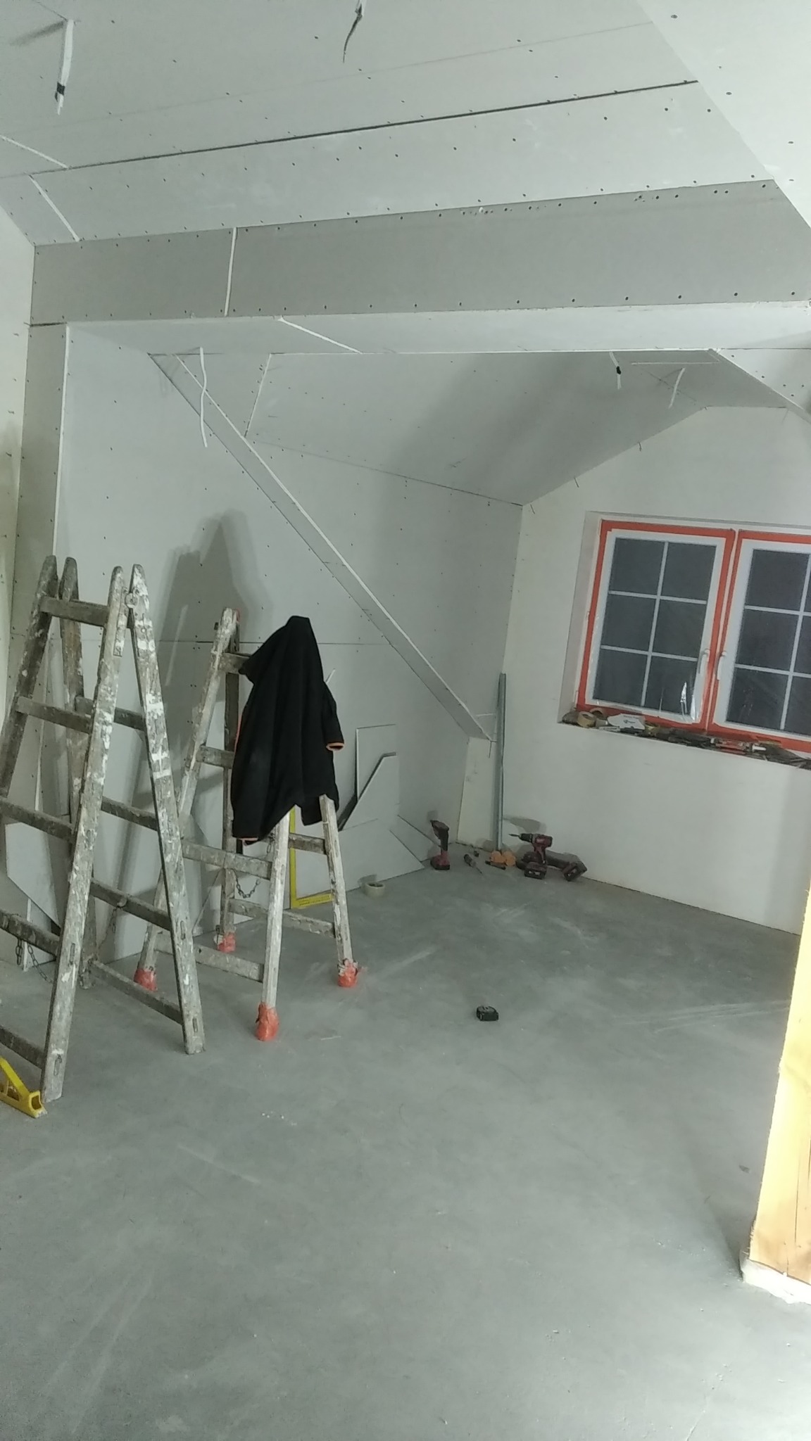 Poddasze w trakcie wykańczania, widoczne płyty gipsowo-kartonowe na ścianach i suficie, dwie drewniane drabiny, narzędzia i czarna kurtka na drabinie. Okno z pomarańczową ramą w tle.
