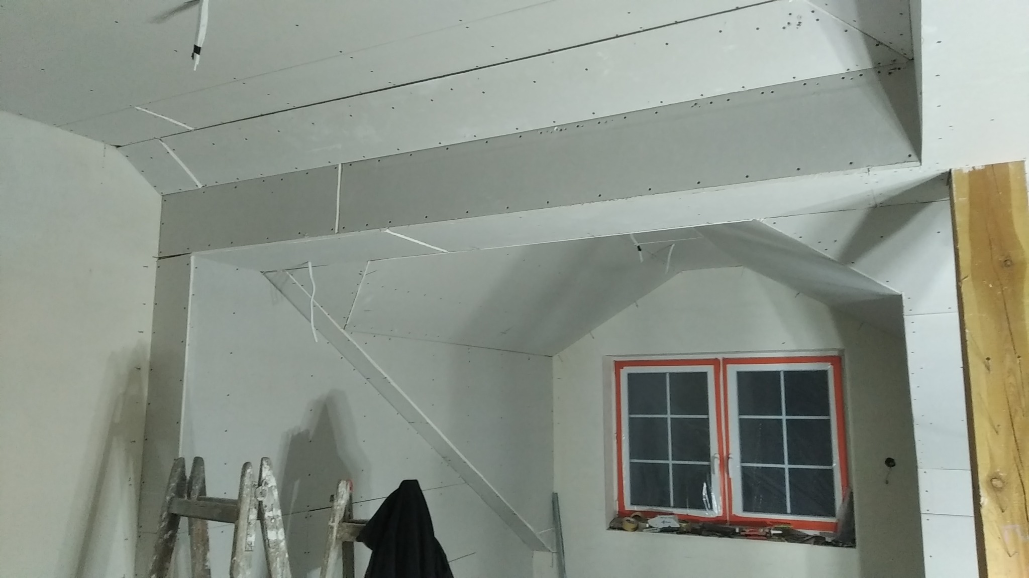 Sufit i ściany w trakcie zabudowy płytami gipsowo-kartonowymi, widoczne łączenia i śruby, okno z pomarańczową ramą w tle, drewniany element konstrukcyjny po prawej.