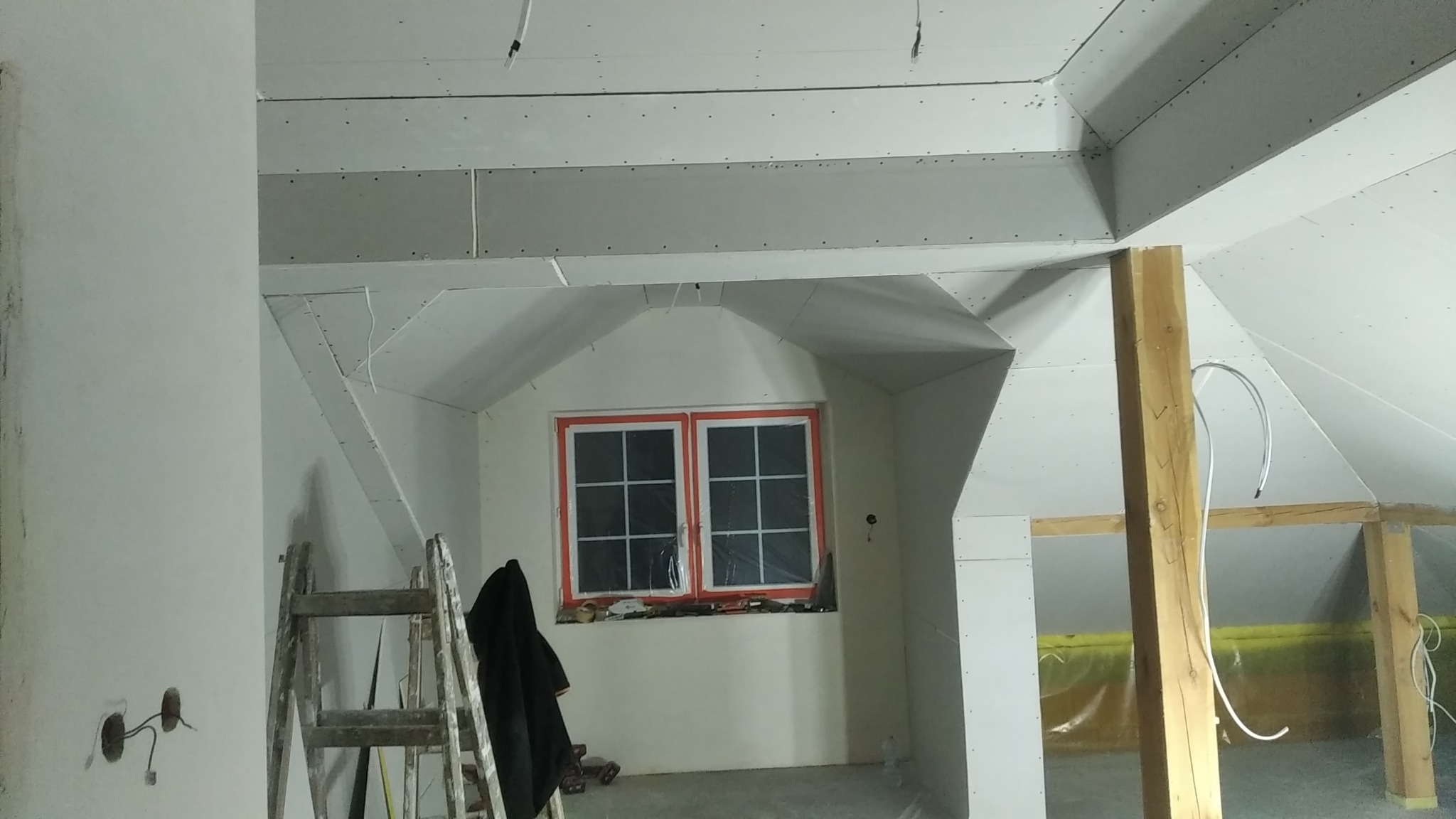 Poddasze w trakcie adaptacji: widoczne płyty gipsowo-kartonowe na skośnych ścianach i suficie, drewniana konstrukcja nośna, drabina oparta o ścianę, okno zabezpieczone folią malarską, instalacja...
