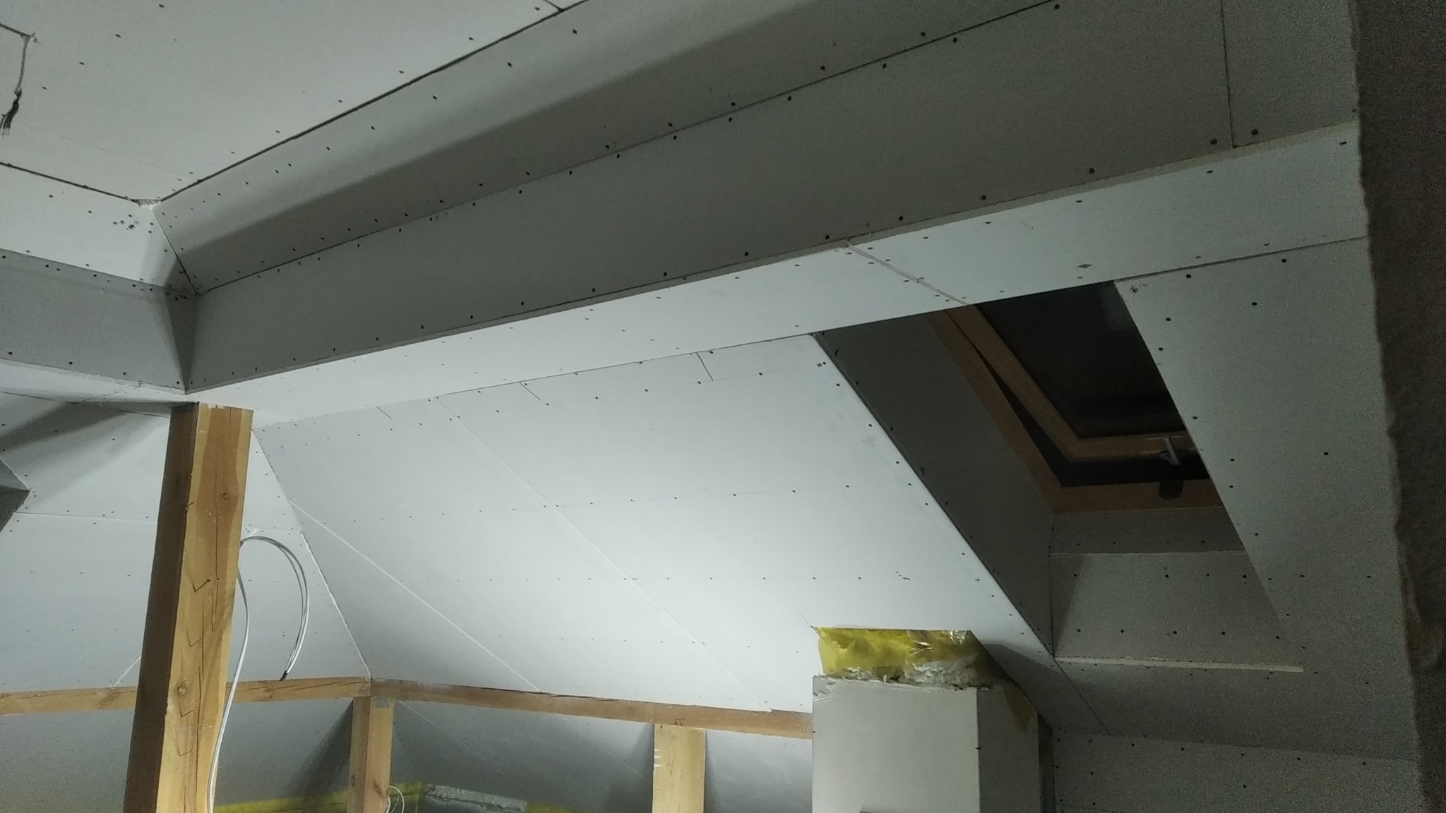 Poddasze w trakcie wykańczania: widoczne płyty kartonowo-gipsowe na skosach sufitu, drewniana konstrukcja szkieletowa oraz fragment okna dachowego.