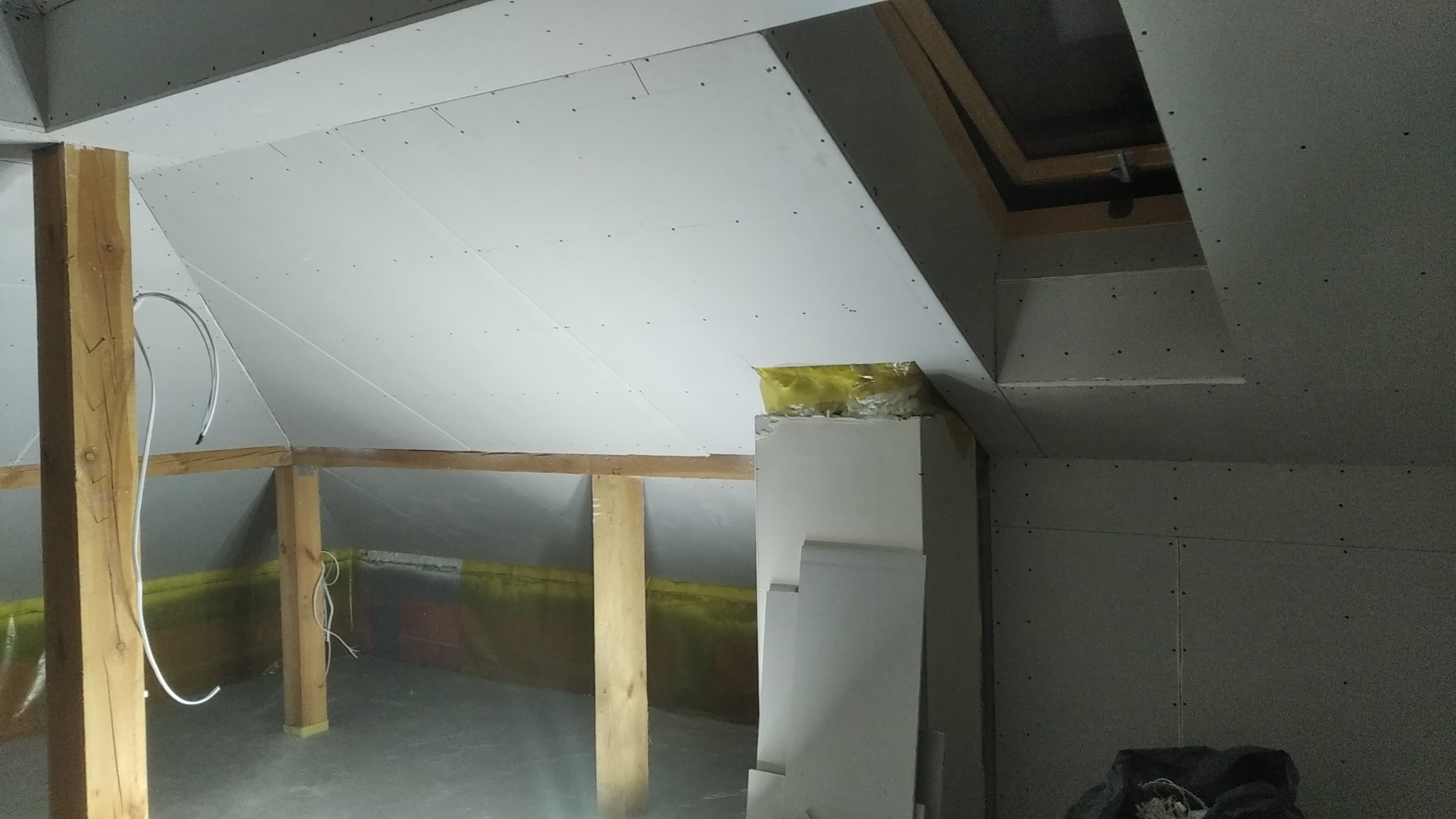 Poddasze w trakcie wykańczania: widoczne drewniane belki, izolacja oraz płyty gipsowo-kartonowe zamontowane na skośnym suficie i ścianach, częściowo zakryte folią ochronną.