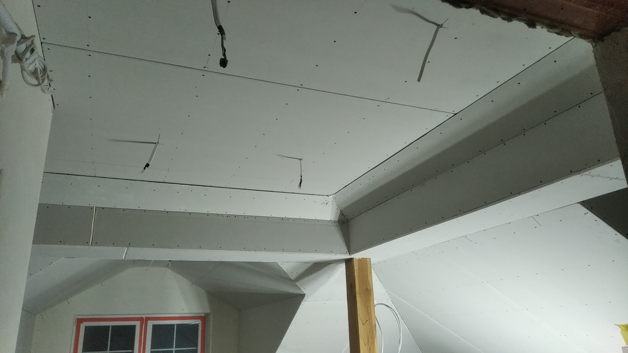 Skośny sufit na poddaszu w trakcie zabudowy płytami gipsowo-kartonowymi; widoczne przewody elektryczne wystające z sufitu i fragment okna w dolnej części kadru.