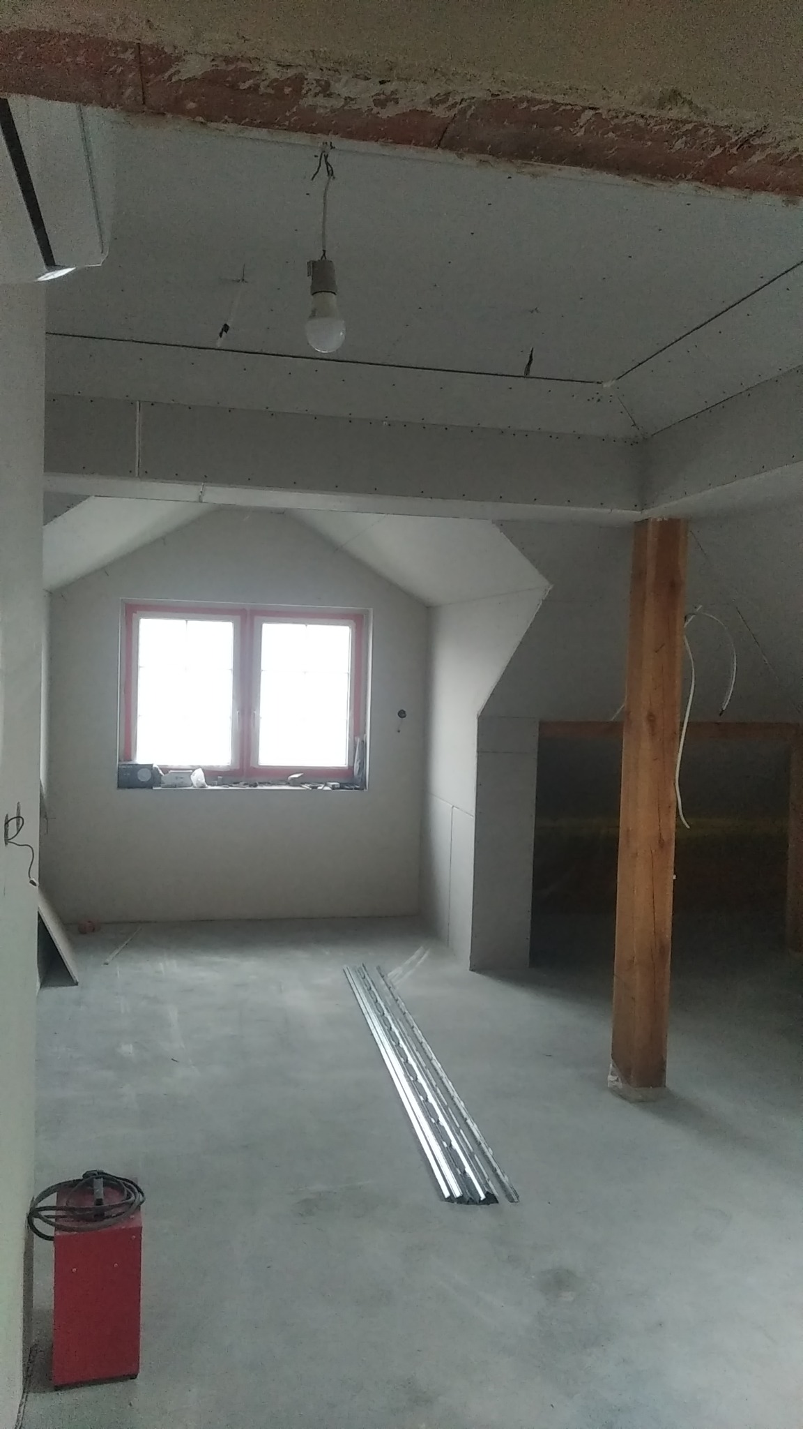 Poddasze w trakcie wykańczania płytami karton gips, widoczne profile na podłodze i czerwony agregat, surowa żarówka zwisa z sufitu.