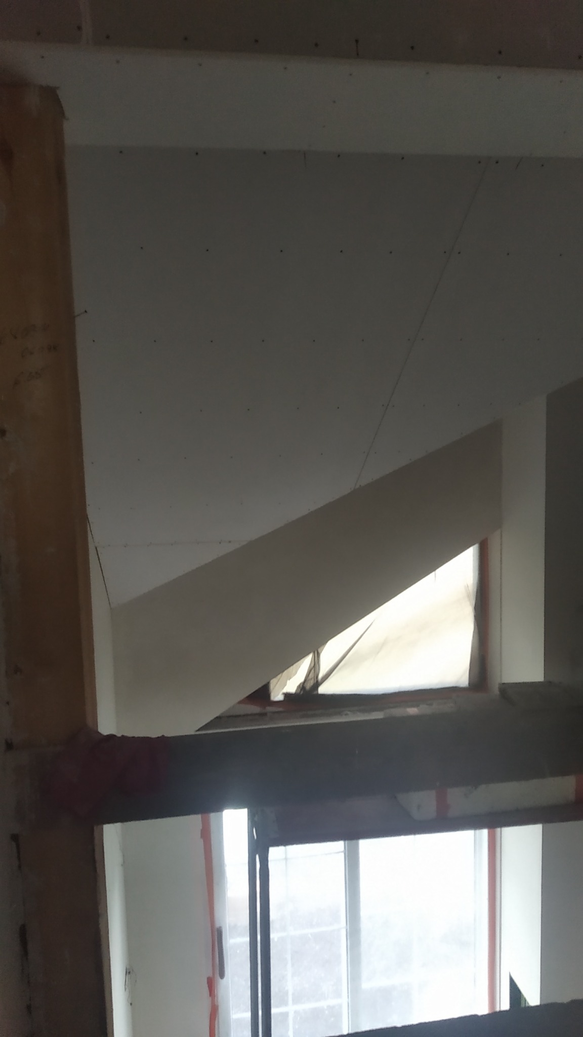 Poddasze w trakcie wykańczania, widoczne płyty gipsowo-kartonowe na skośnym suficie i ścianie, drewniana belka konstrukcyjna oraz okno zasłonięte folią ochronną.