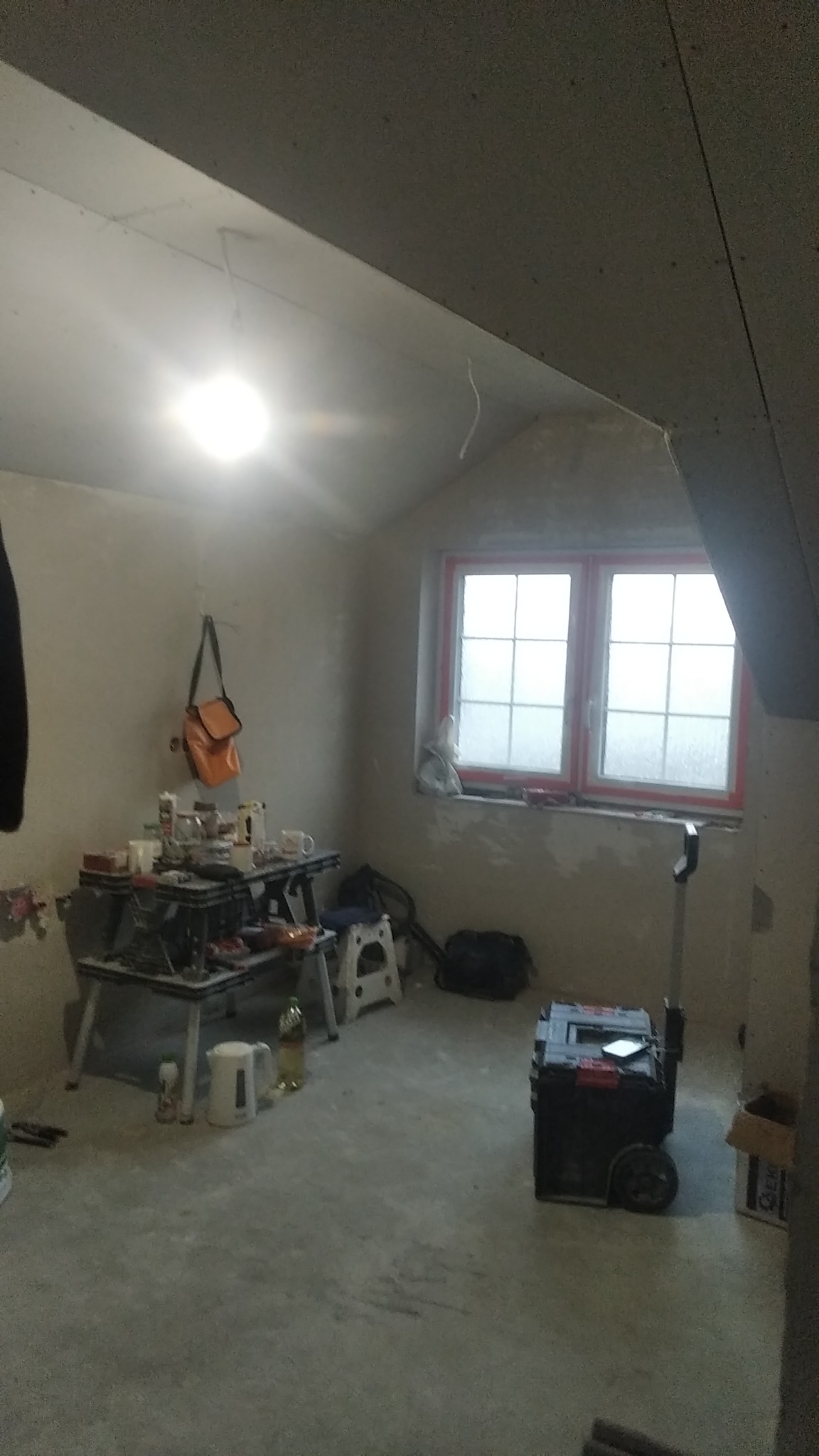 Poddasze w trakcie prac wykończeniowych, widoczne płyty na skosach sufitu, narzędzia i materiały rozłożone na prowizorycznym stole roboczym, okno z czerwoną ramą.
