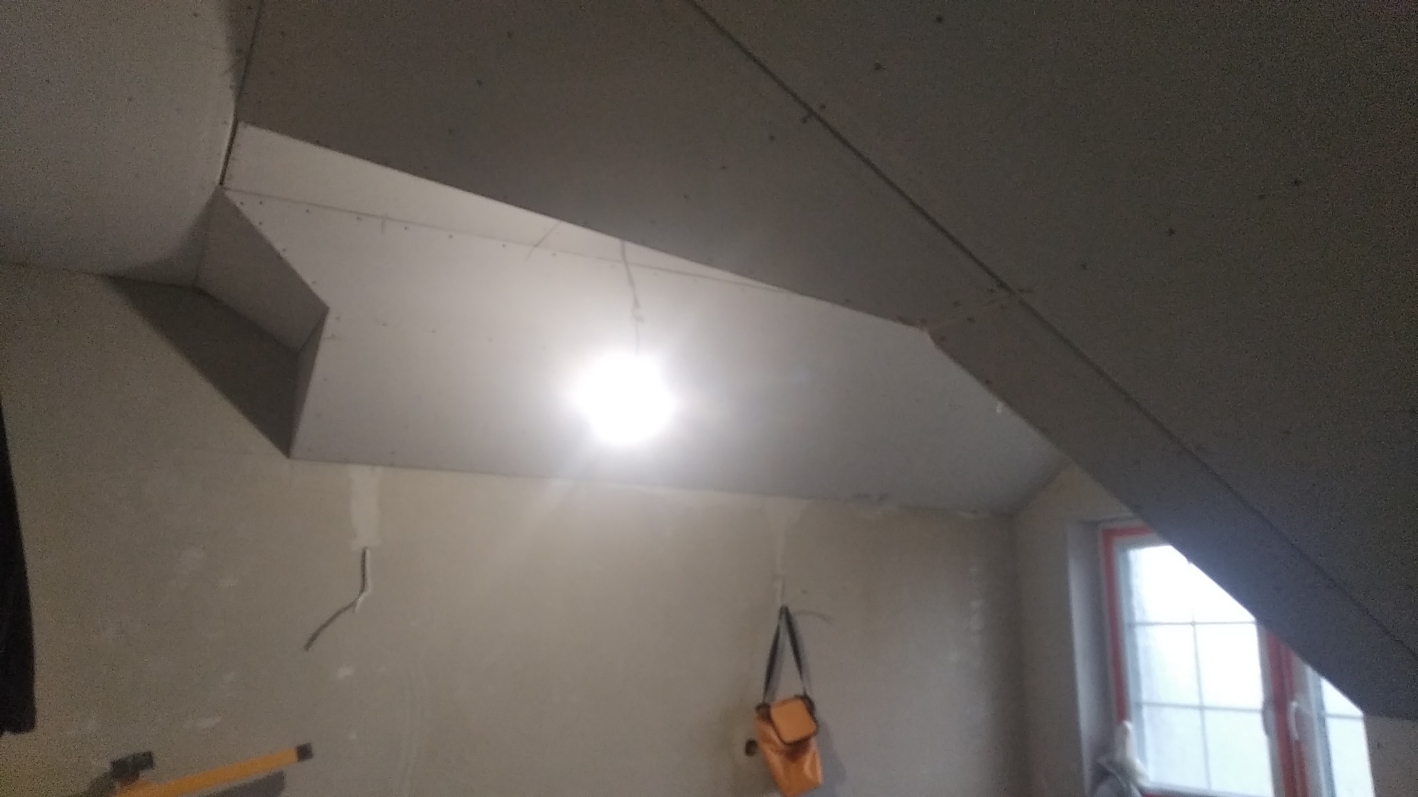 Montaż płyt na skośnym poddaszu, widoczna konstrukcja sufitu, wisząca torba narzędziowa i fragment okna w tle.