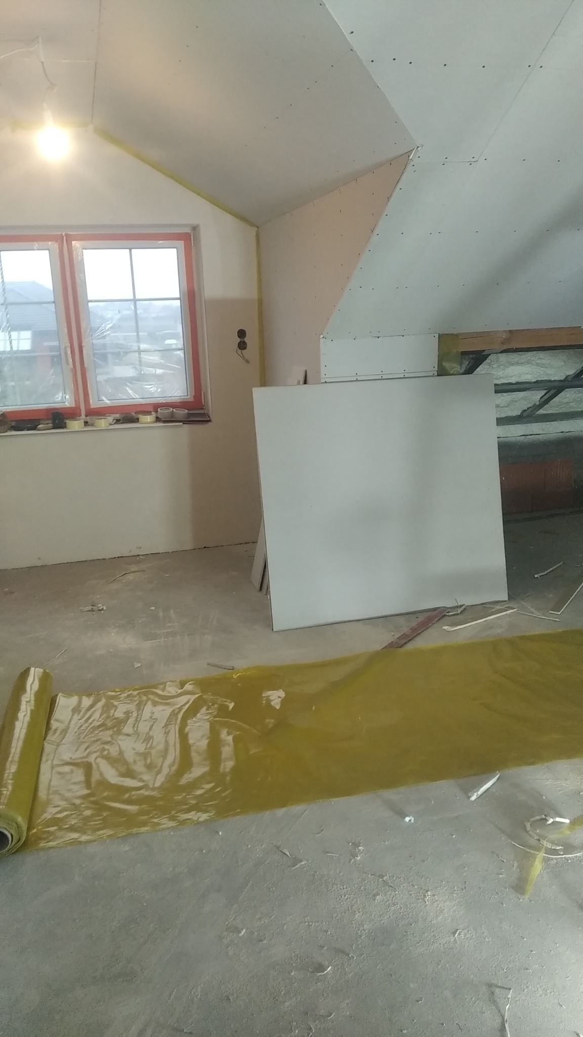 Poddasze w trakcie wykańczania, widoczne płyty gipsowo-kartonowe na suficie i ścianach, okno z pomarańczową ramą, folia ochronna na podłodze i niezagruntowane ściany.