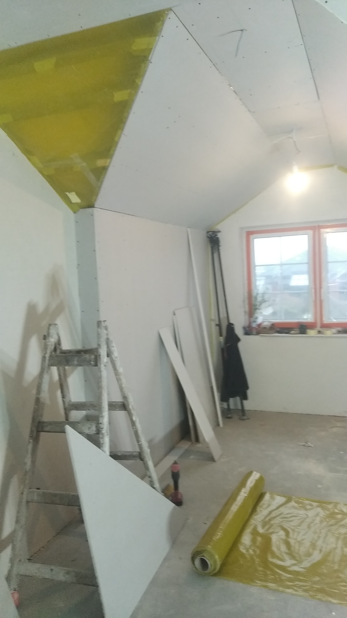 Poddasze w trakcie wykańczania: częściowo zamontowane płyty kartonowo-gipsowe na suficie skośnym, widoczna drabina, rolka folii ochronnej i narzędzia.