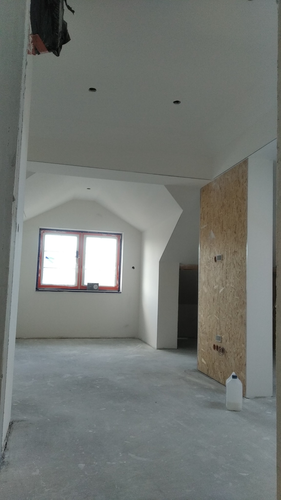 Pokój na poddaszu w trakcie wykańczania, widoczne ściany pokryte gładzią, okno dachowe z pomarańczową ramą oraz fragment ściany z płyty OSB z puszkami elektrycznymi.