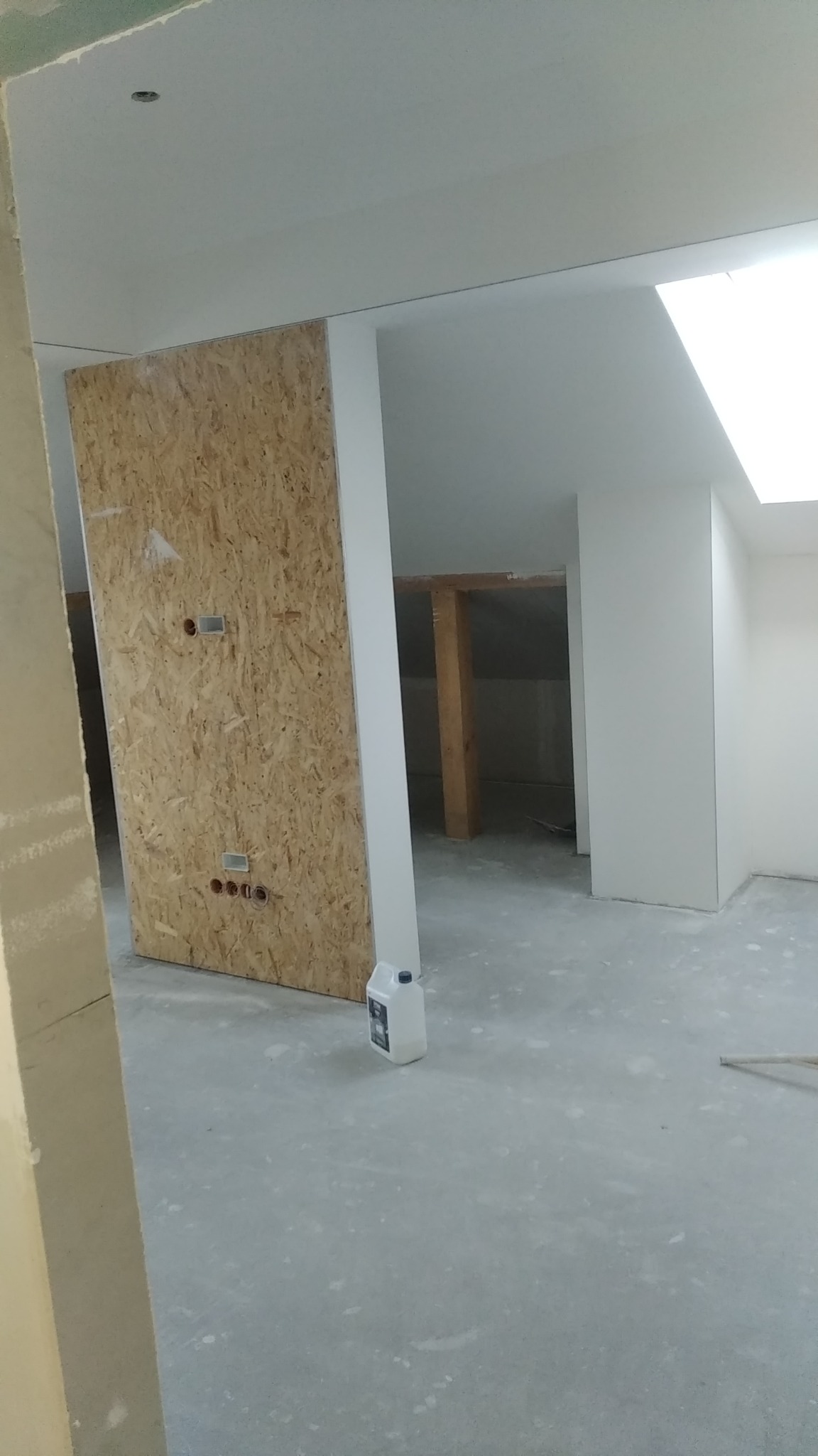 Płyta OSB zamontowana na ścianie z wyciętymi otworami na gniazdka elektryczne w surowym wnętrzu z oknem dachowym, widoczna butelka z preparatem do gruntowania podłoża.