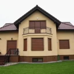 Elewacja domu jednorodzinnego z zamkniętymi roletami zewnętrznymi w kolorze ciemnego drewna, balkon z ozdobną balustradą, brązowy dach z dachówki.