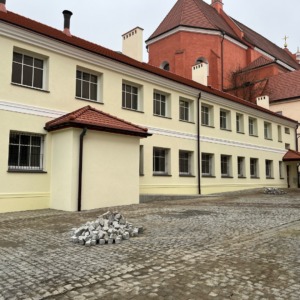 Elewacja budynku z jasnymi oknami z podziałami, widoczna sterta kostki brukowej na dziedzińcu i fragment czerwonego budynku w tle.