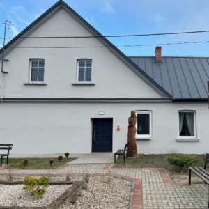 Elewacja budynku z szarymi oknami PCV i ciemnymi drzwiami wejściowymi, widok z przodu, na pierwszym planie ścieżka z kostki brukowej i rabaty kwiatowe, po prawej drewniana rzeźba.