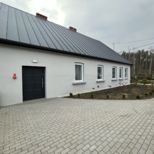 Budynek z szarym dachem, jasną elewacją, ciemnymi drzwiami wejściowymi i kilkoma białymi oknami. Ułożona kostka brukowa na podjeździe.