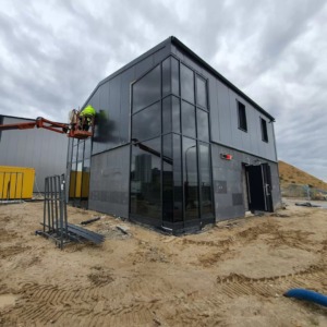 Montaż okien w nowoczesnym, dwukondygnacyjnym budynku z szarej blachy trapezowej. Pracownik na podnośniku koszowym instaluje duże, czarne okno. W tle widoczny teren budowy z piaskiem i kontenerami.