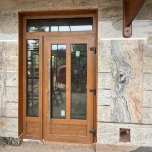 Zewnętrzne drzwi wejściowe z drewna w odcieniu miodowym, z bocznym panelem szklanym i nadświetlem, osadzone w elewacji z jasnego granitu z wyraźnym wzorem żyłkowania.